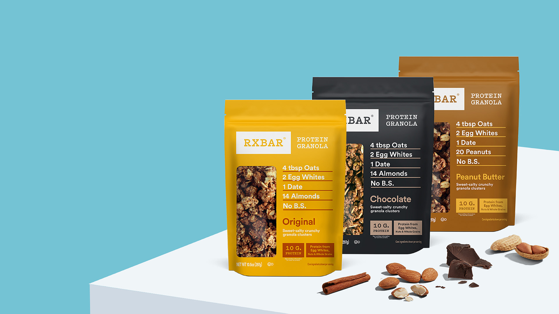 Pouches of original, chocolate, and peanut butter RXBAR Granola, on a table with a cinnamon stick, nuts, and chocolate chunks
